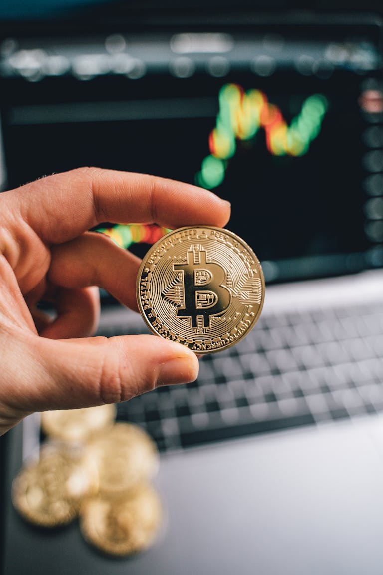 A hand holds a Bitcoin coin against a laptop with a trading graph display, symbolizing cryptocurrency trade.